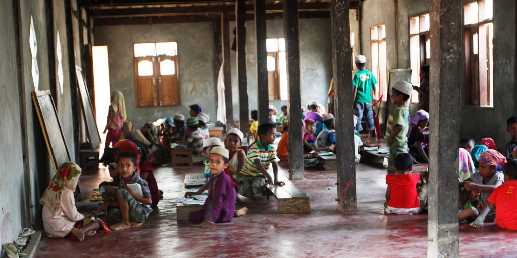 Rohingya children sitting on a red concrete floor in a sparsely furnished room. Some are looking toward the camera and others are engaged in school lessons.