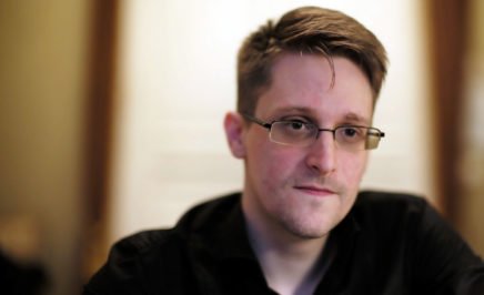 A close up shot of whistleblower Edward Snowden, wearing glasses and a black shirt, staring into the camera