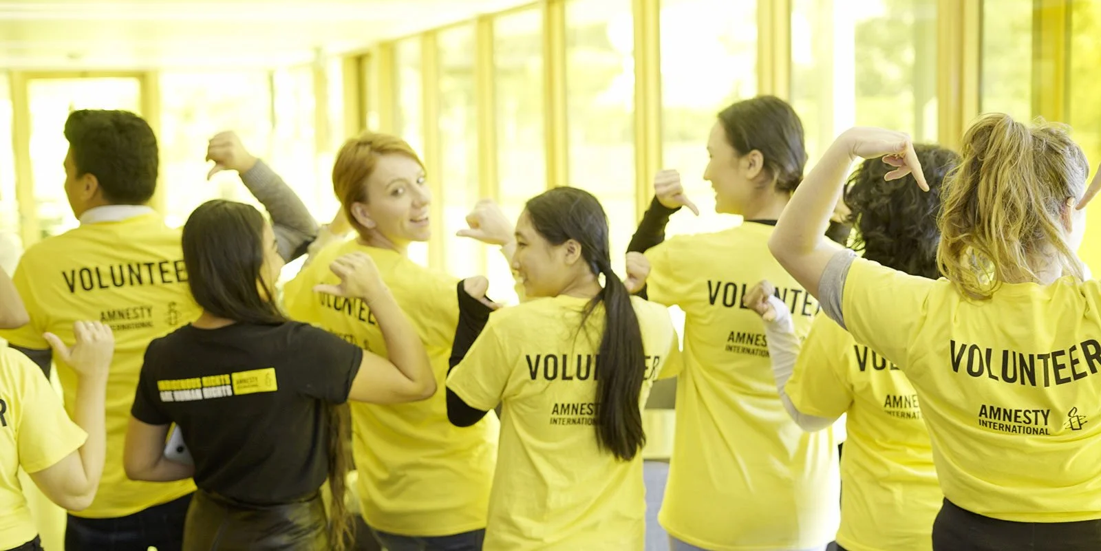 A photo of a group of eight Amnesty volunteers (both men and women) wearing Amnesty tshirts. The volunteers have their backs to the camera and the word 'Volunteer' is emblazoned across the back of their tshirts.