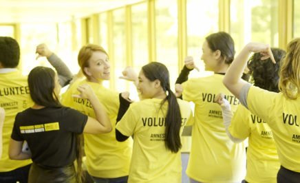 A photo of a group of eight Amnesty volunteers (both men and women) wearing Amnesty tshirts. The volunteers have their backs to the camera and the word 'Volunteer' is emblazoned across the back of their tshirts.