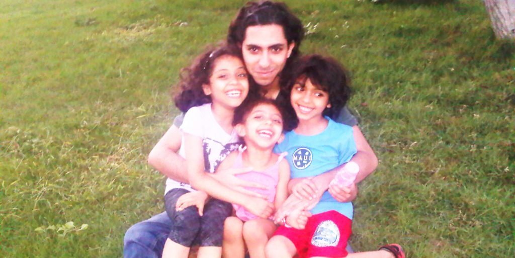 A man sits of a patch of grass with his three children: two girls and a boy. All four are smiling for the camera.
