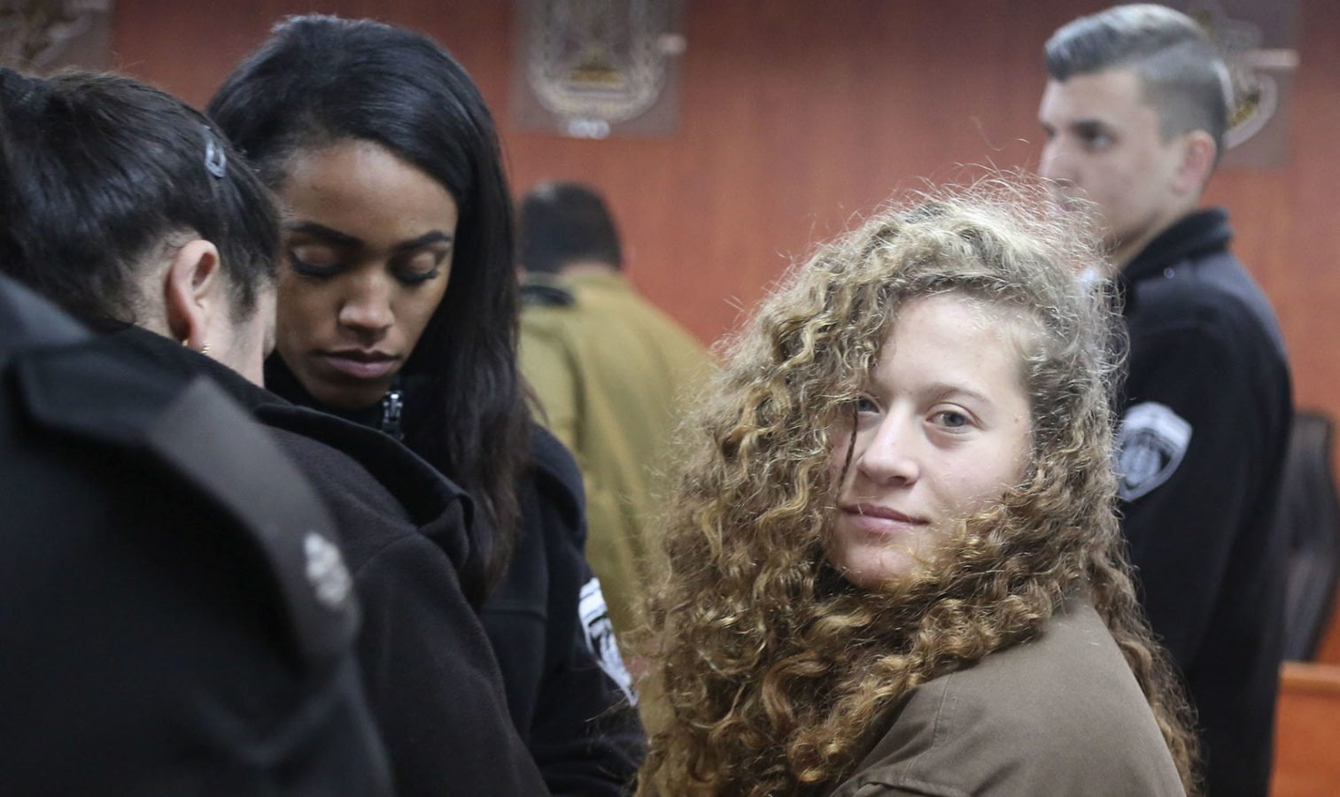 A young woman with curly hair and wearing a brown jacket stands in a court room surrounded by guards. She is looking at the camera.