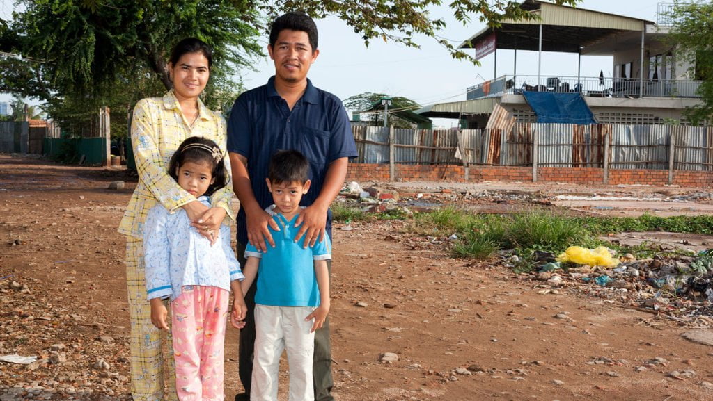 Cambodian rights activist Tep Vanny standing with her husband and two child, a boy and a girl, smiling at the camera. The family are outside, standing on dried, muddy ground, with a housing structure in the background.