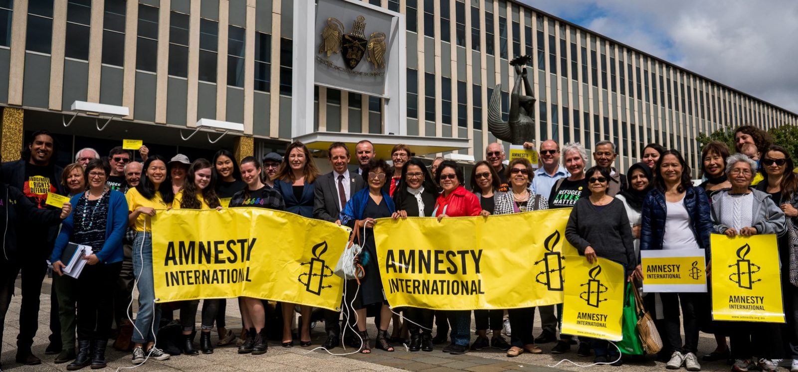 A group of people holding yellow Amnesty banners standing outside Parliament House in Canberra