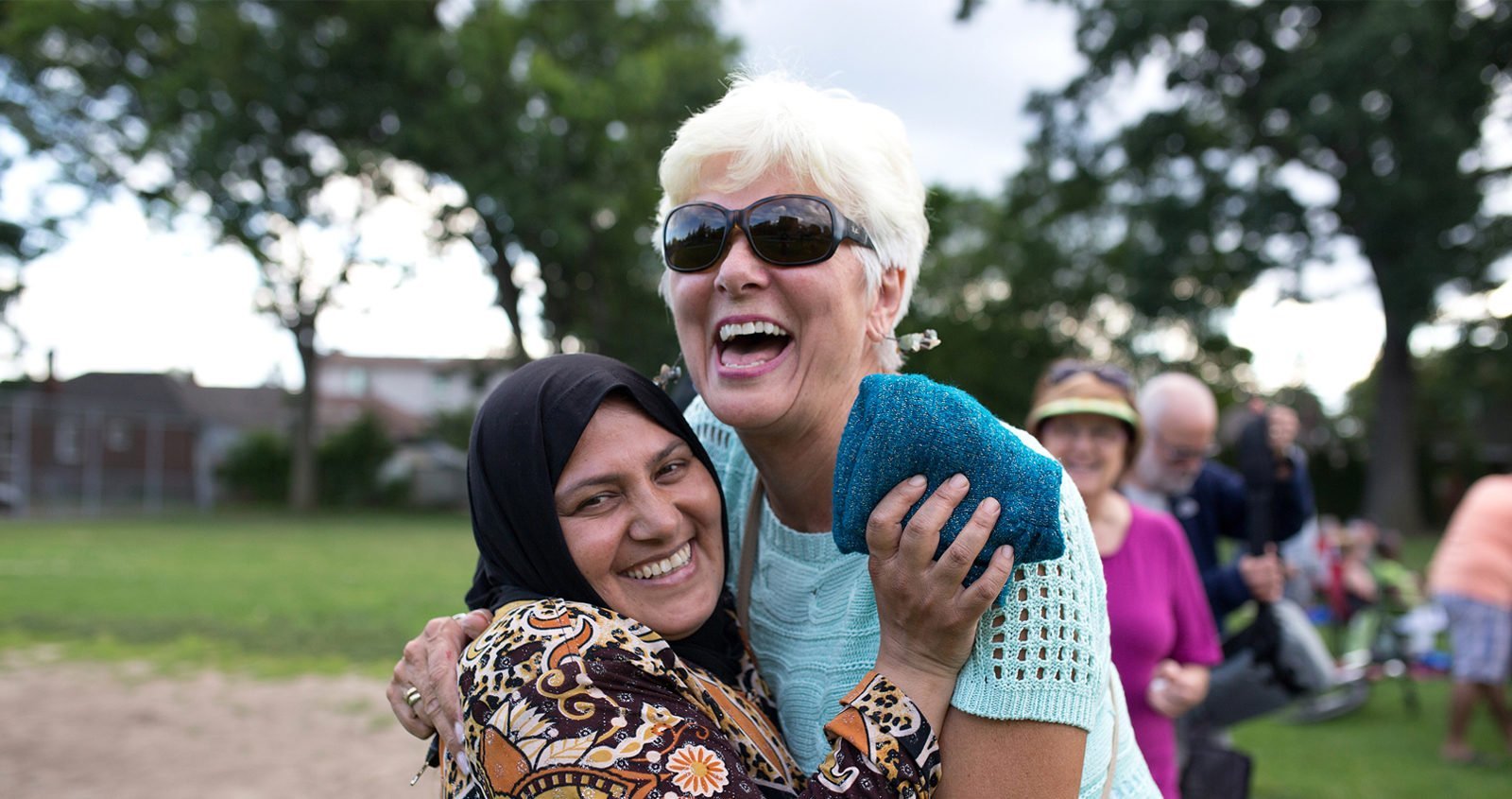 Woman in with head scarf hugs a laughing, middle-aged woman of Western appearance.