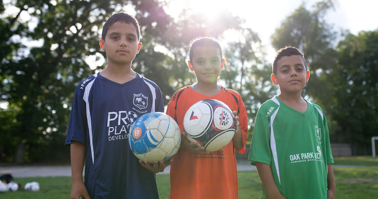 Three children holding soccer balls and wearing soccer jerseys in a sunlit park.