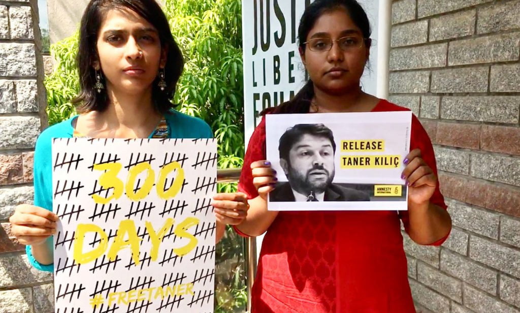 Two Indian women standing side by side. One is holding a sign of a tally of the number of days Taner Kilic has spent in jail with the words '300 days, #FreeTaner' on it and one holding a sign with a photo of Taner on it that says 'Release Taner Kilic'