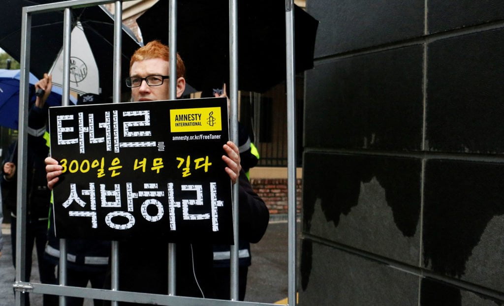 Man holding a sign behind fake bars, protesting the imprisonment of Taner Kılıç