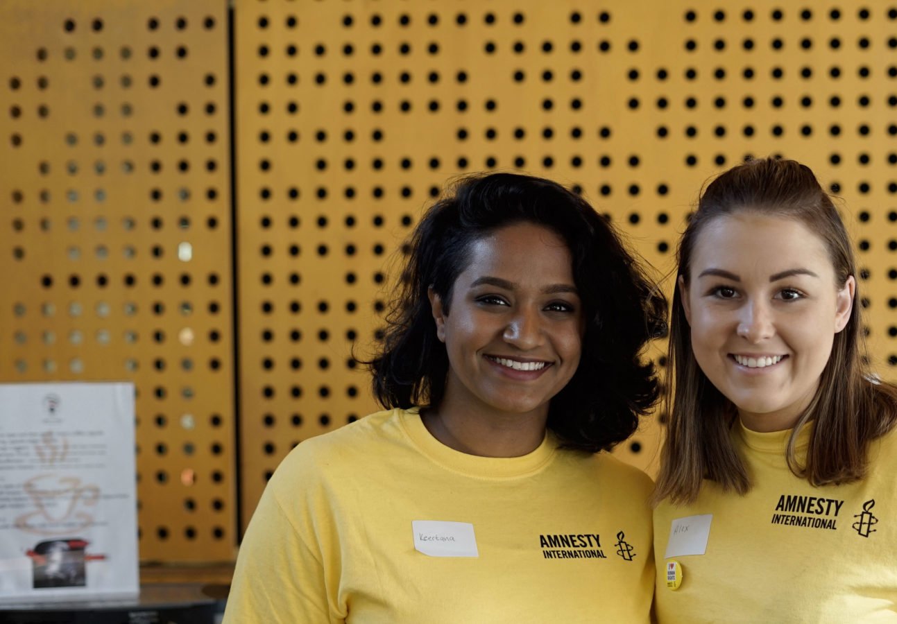 Two female Amnesty volunteers wearing yellow t-shirts smile at the camera.