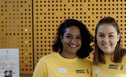 Two female Amnesty volunteers wearing yellow t-shirts smile at the camera.