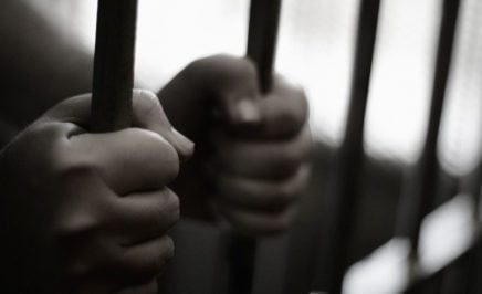 Two hands holding onto a set of prison bars in a jail cell