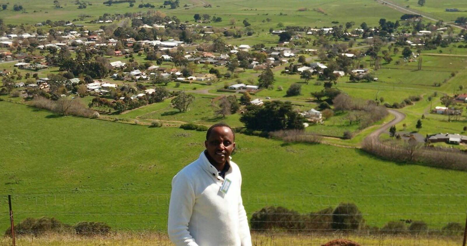 Man with dark skin in business attire with green, rural farmlands behind him.