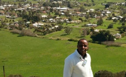Man with dark skin in business attire with green, rural farmlands behind him.
