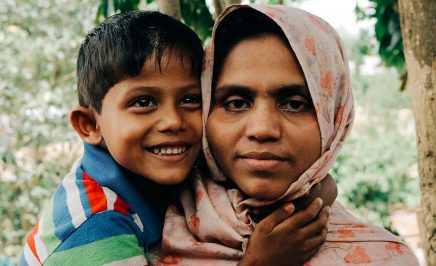 A Rohingya woman holds her child. The woman is looking straight into the camera and the child is smiling and looking off-camera.
