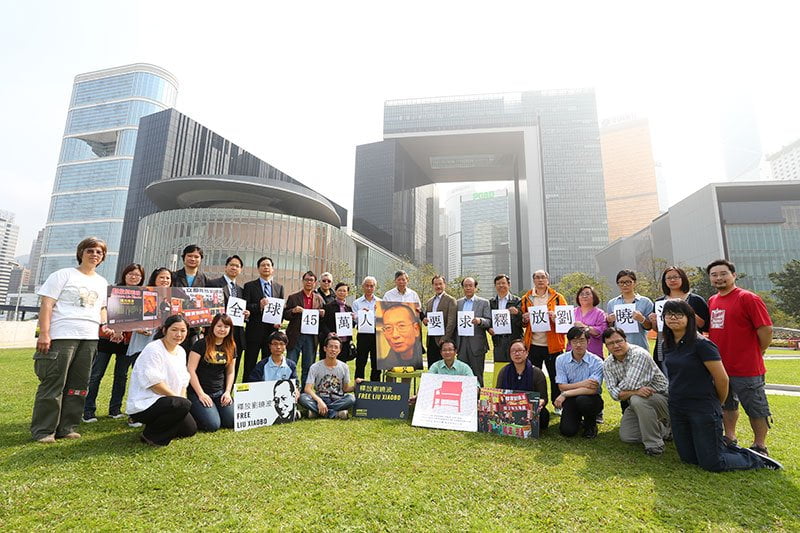 A group of people standing on grass in front of large glass office buildings in Hong Kong and holding signs and images calling for Liu Xia and Liu Xiaobo to be freed.