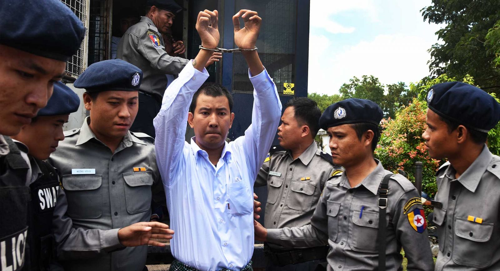 A man is walking with his hands cuffed above his head. He is flanked by six members of the Myanmar police force.