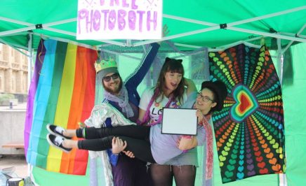 three people dressed in bright colours hug under a sign saying free photobooth