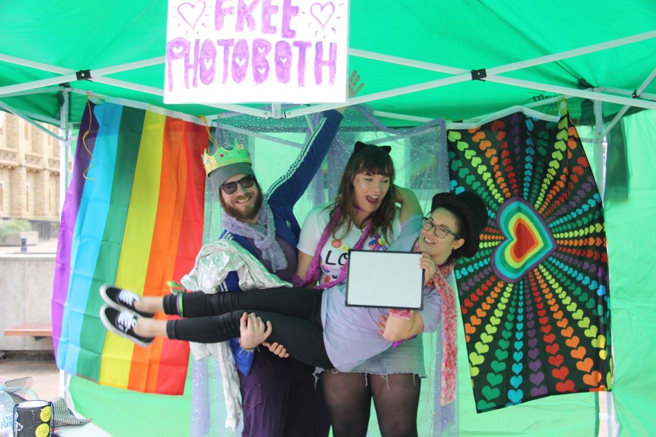 three people dressed in bright colours hug under a sign saying free photobooth