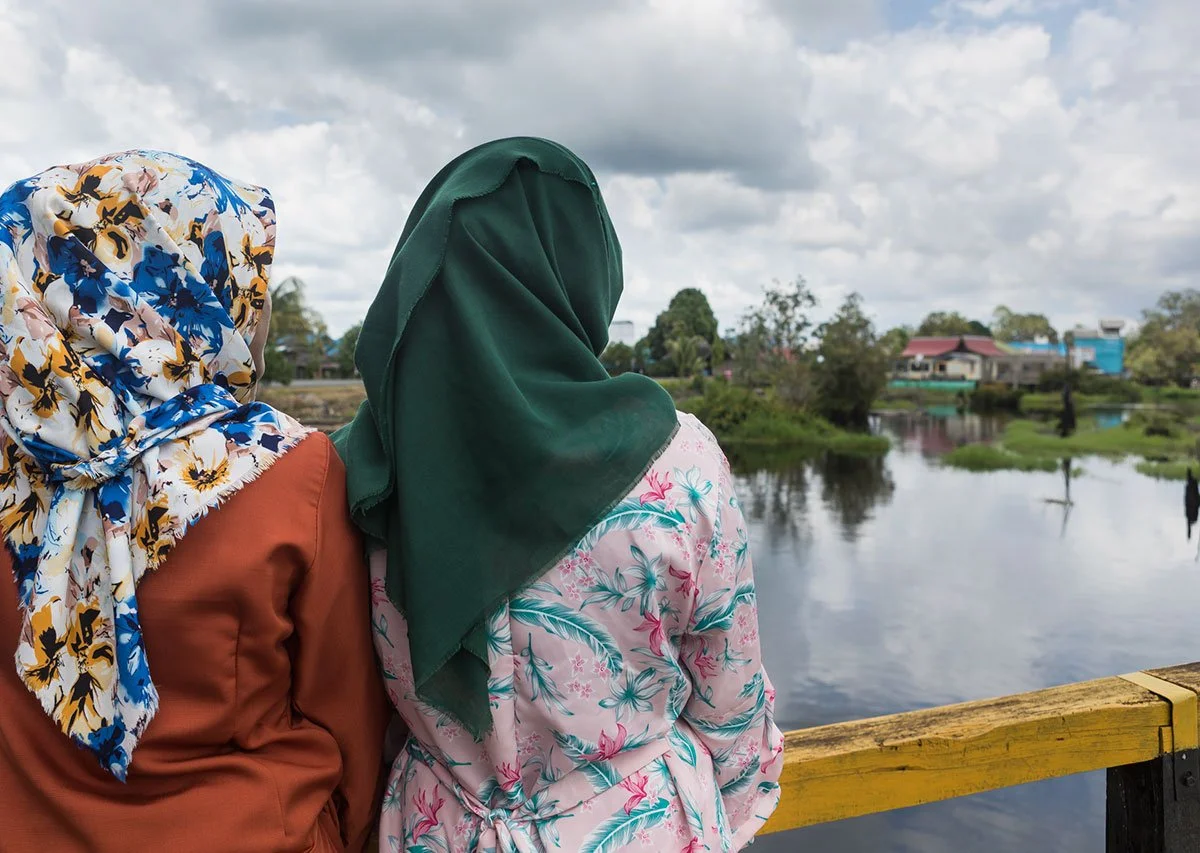 An image of three Indonesian women staring out over a river towards a village in the distance. The women have their backs to the camera and are wearing brightly coloured head scarves.