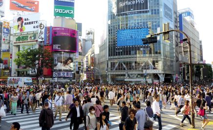 An image of a bustling street in Tokyo, Japan. Hundreds of people cross an intersection and tall buildings with brightly lit screen line the street.