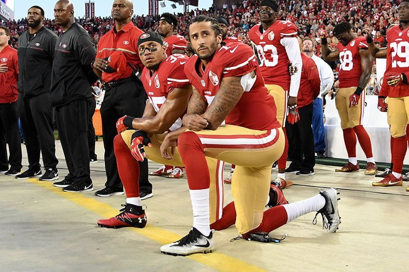 Two men dressed in the red and yellow team strip of the San Francisco 49ers kneel in protest during the singing of the American national anthem before a football game. Behind them their team mates are standing and in the ba