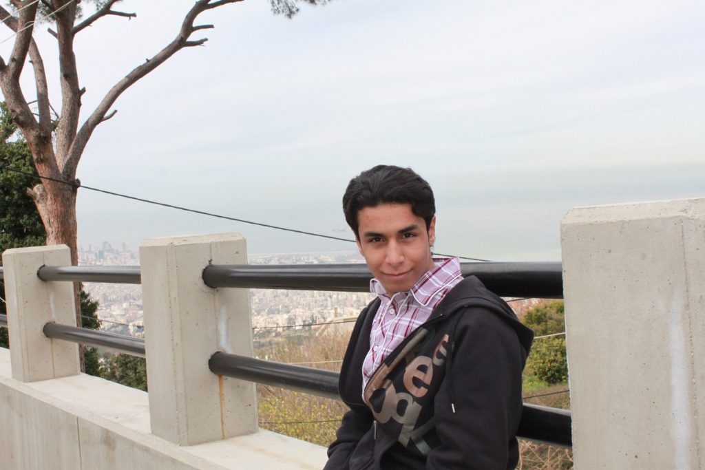 A young man from Saudi Arabia smiles at the camera. He's wearing a checked shirt and a hoodie. A city can be seen in the background.