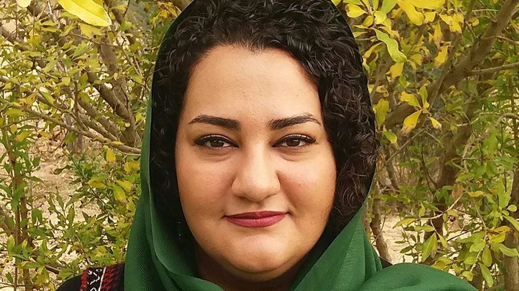 A woman wearing a green headscarf smiles at the camera. She is set against a leafy backdrop of trees.