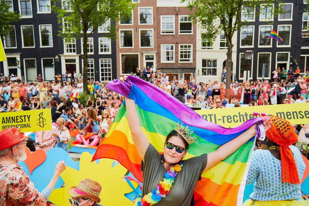 A woman wearing sunglasses stands at the front of a crowd at the Canal Parade in Amsterdam. She is holding an LGBTQI rainbow banner.