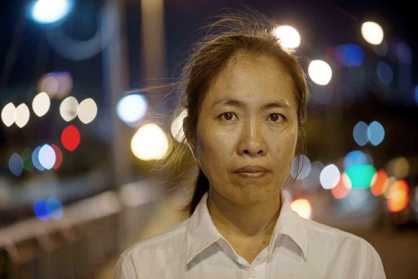 A Vietnamese women wearing a white shirt stares into the camera. It is nighttime and the lights of a city twinkle behind her.
