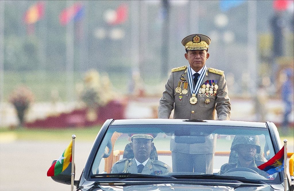 A man in a Myanmar military uniform stands up in an open-top car at a military parade. There are two other people in the car with him, also in military dress. Blurred in the background we can see a line of flags from different countries.