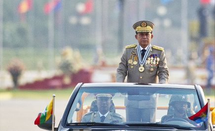 A man in a Myanmar military uniform stands up in an open-top car at a military parade. There are two other people in the car with him, also in military dress. Blurred in the background we can see a line of flags from different countries.