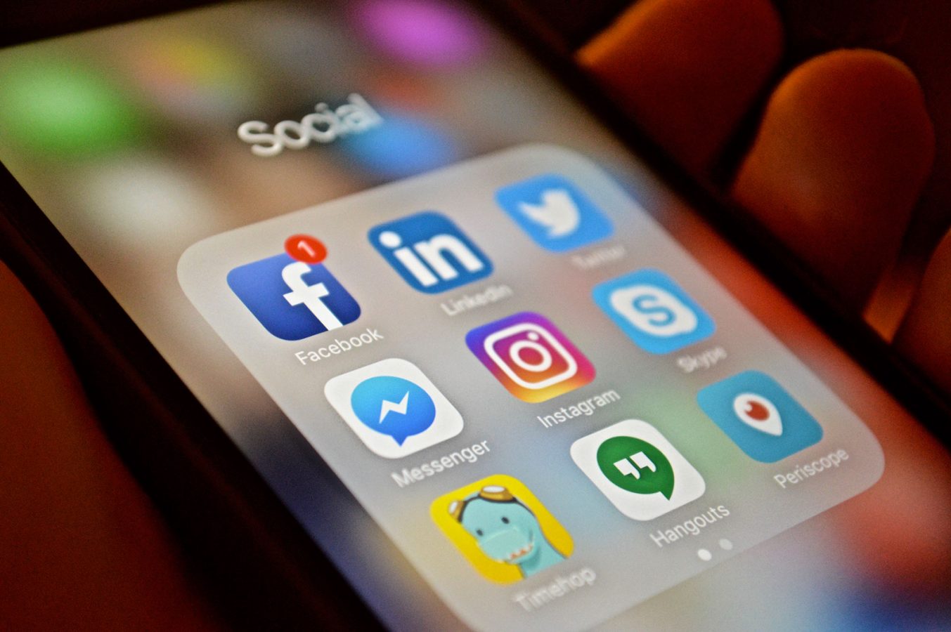 A hand holding a mobile phone displaying the icons of social media apps: Facebook, LinkedIn, Twitter, Messenger, Instagram, Skype, Timehop, Hangouts and Periscope.