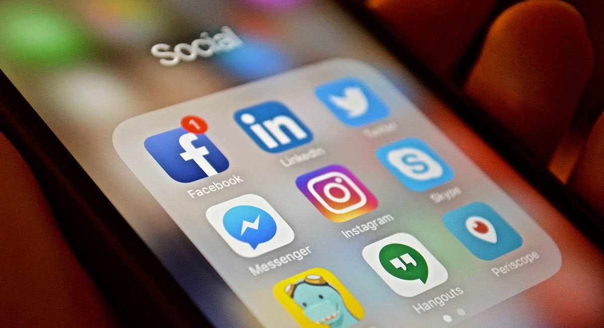A hand holding a mobile phone displaying the icons of social media apps: Facebook, LinkedIn, Twitter, Messenger, Instagram, Skype, Timehop, Hangouts and Periscope.