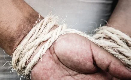 A photo of a man's hands bound behind his back with fraying twine or rope.