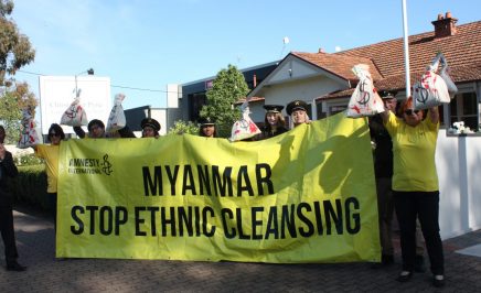 a group of people standing on a footpath with a large yellow sign saying 'Myanmar stop ethnic cleansing'