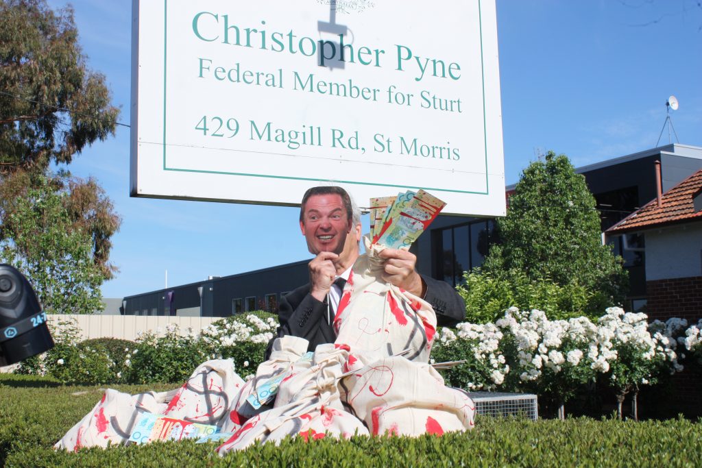 man sits in front of sign saying 'christopher pyne federal member for sturt' with a paper mask, holding bags of money