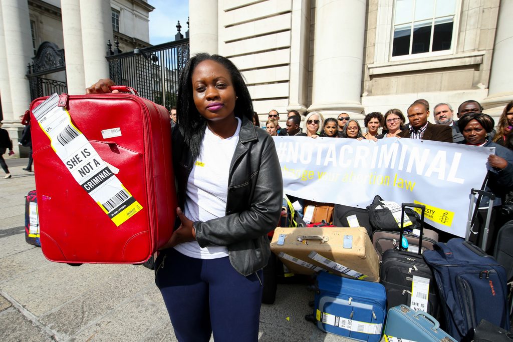 A woman stands in front of the Irish Taoiseach's offices in Dublin holding a suitcase with a boarding sticker with 'She is not a criminal' printed on it. Behind her stand a group of activists holding a banner that says the same thing.