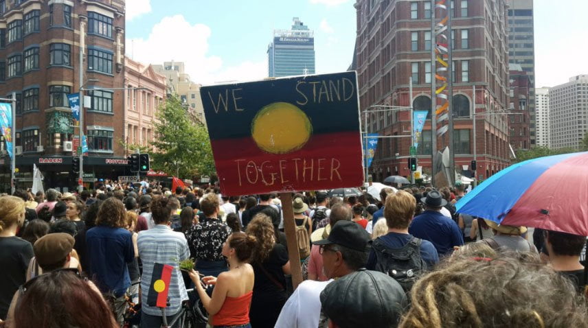 People gather to protest for Indigenous rights. Someone holds a sign that says: 'We stand together'.