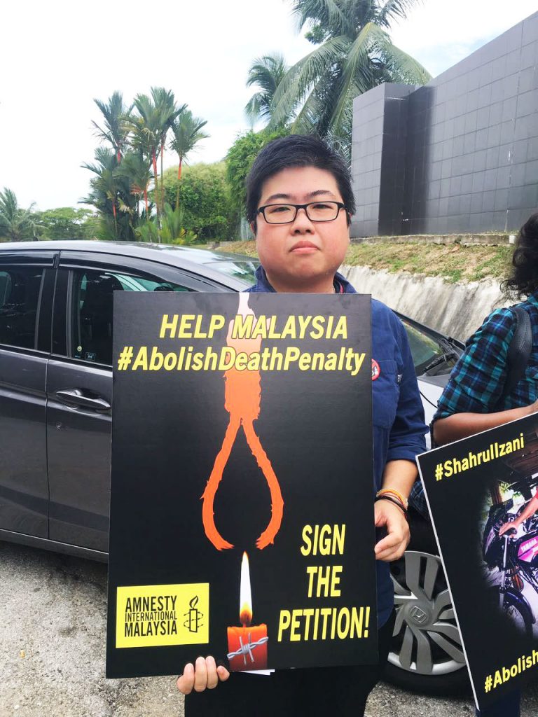 A Malaysian man holds a sign towards the camera. The sign reads: 'Help Malaysia #AbolishDeathPenalty