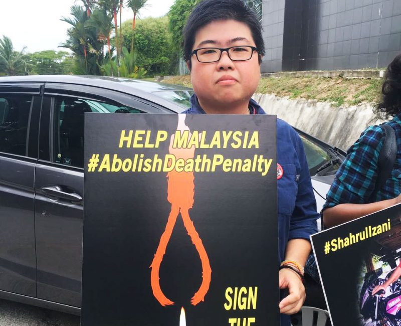A Malaysian man holds a sign towards the camera. The sign reads: 'Help Malaysia #AbolishDeathPenalty