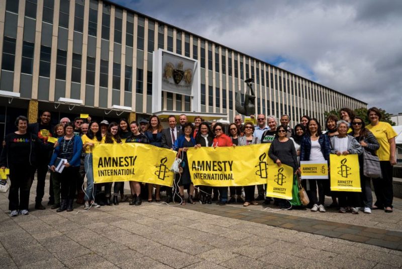 A crowd of about 30 people outside Canberra parliament, holding yellow Amnesty banners