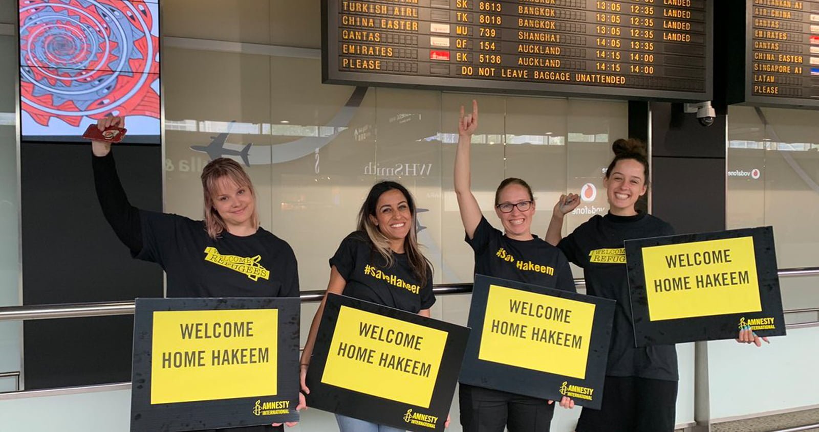 a group of supporters wearing #SaveHakeem shirts and holding 'welcome home Hakeem' signs under a flight arrivals board
