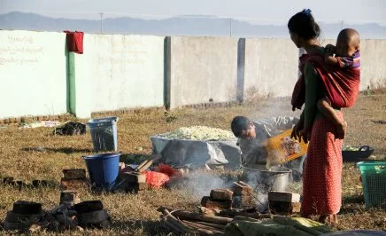 A woman with a child strapped to her back stands on grass watches as a man pours water for cooking rice into a large pan. In the background is a wall and living utensils are scattered around them on the grass.