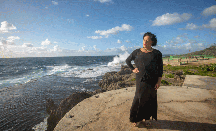 A woman in a long black dress stands on a cliff next to the ocean in Tonga and stares into the distance.