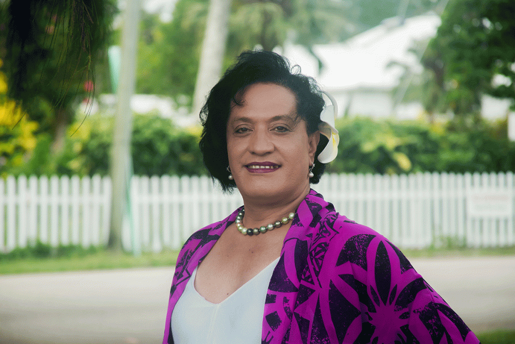 A woman wearing a white top with a purple jacket smiles into the camera. In the background is a neighbourhood street with trees and white picket fences.