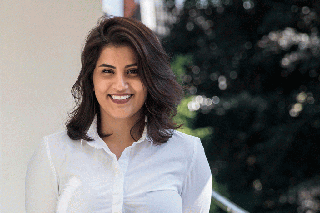 A young woman with dark hair and wearing a white shirt smiles at the camera. Tree foliage is visible in the background.