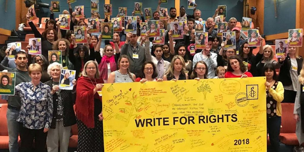 a big crowd of people hold up a yellow banner that says Write for Rights