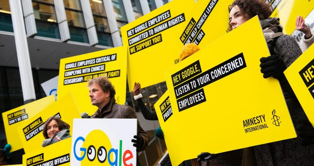 A group of people holding very bright yellow placards, with writing: Hey google, listen to your concerned employees. Hey google employees committed to human rights: we support you!