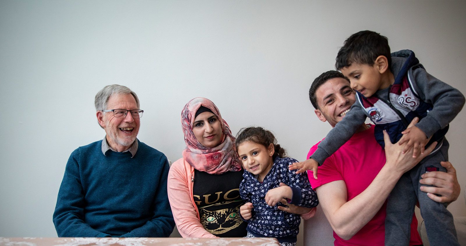 A family from Syria - two parents and two children - with an elderly gentleman, sitting around a table laughing as the boy climbs onto his dad's lap.