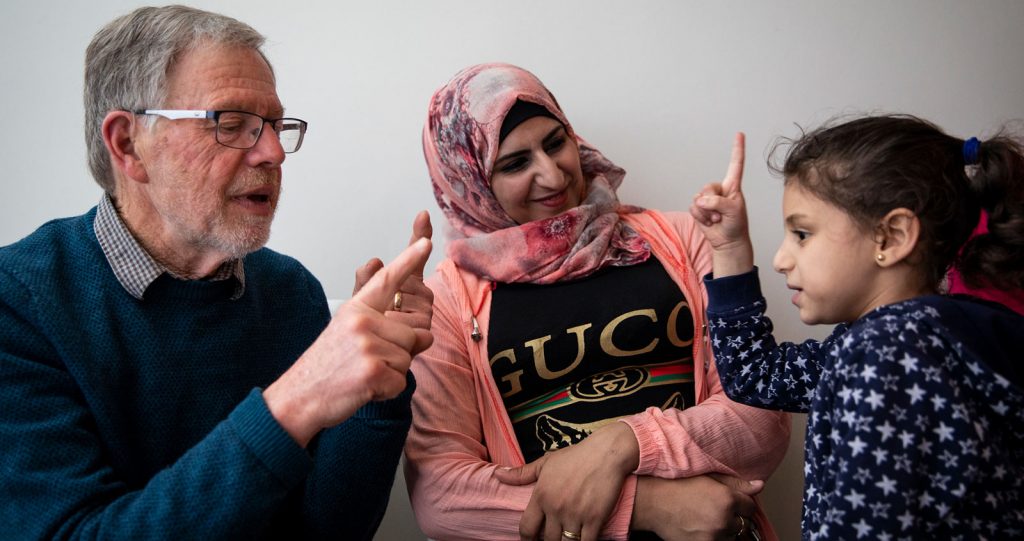 An elderly man gestures at a young girl as if he is teaching her how to count. The girl's mother sitting between them, smiling.